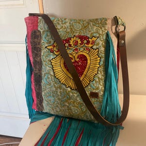 K&ouml;nnte beinhalten: Eine t&uuml;rkisfarbene und rote Umh&auml;ngetasche mit Fransen und einem braunen Lederriemen. Die Tasche hat ein gro&szlig;es gesticktes Herz mit einer Krone und Pailletten.
