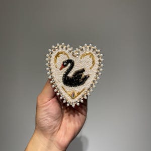 May include: Heart-shaped brooch with a black swan design. The brooch features a cream-colored background, gold accents, and is embellished with small pearls and crystals. The swan has a red beak.