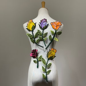 Può includere: Cinque applicazioni ricamate a forma di rosa in vari colori, tra cui rosa, giallo, arancione e viola, con steli e foglie verdi. Le applicazioni sono disposte su un manichino bianco.