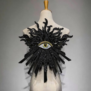 May include: A black, sunburst-shaped accessory with a central eye design. The accessory is adorned with small, sparkling embellishments. The eye features a blue iris, a yellow iris ring, and a white sclera. The accessory is displayed on a mannequin.