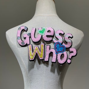 May include: Pink fabric patch with the words "Guess Who?" in sparkly sequins. The letters are outlined in black and have different colored sequins.