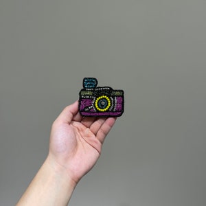 May include: A beaded camera brooch with a purple, black, and yellow design. The camera has a small black flash on top.