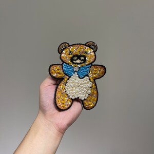 May include: A brown teddy bear applique with yellow sequins, white pearl beads, and a blue bow tie.