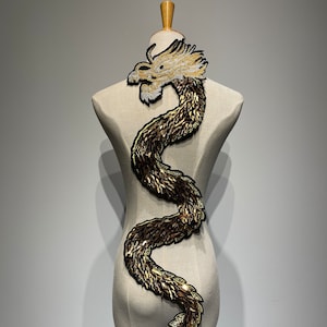 May include: Gold and white embroidered dragon applique. The dragon is curled in a serpentine shape with its head facing to the left. The applique is perfect for adding a touch of elegance to clothing, bags, or home decor.