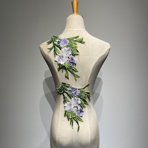 May include: Embroidered floral appliques featuring clusters of lavender and white flowers with green leaves. The appliques are displayed on a mannequin, showcasing the design and potential placement for garment decoration or craft projects.