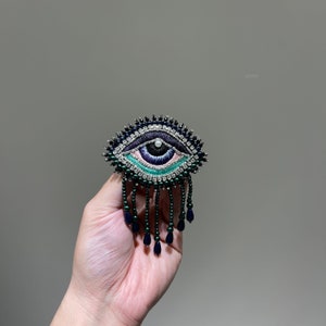 May include: A black and green eye-shaped brooch with a beaded fringe. The eye is embroidered with blue, green, and pink thread and has a white pearl in the centre.