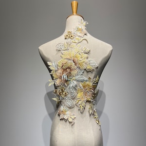 Può includere: Applicazione floreale ricamata nei toni del giallo, marrone e argento, esposta su un manichino. Il design presenta fiori e foglie dettagliati, creando un effetto strutturato e tridimensionale. L'applicazione è probabilmente per abbellire capi d'abbigliamento o lavori artigianali.