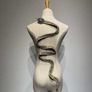 Può includere: Un design a forma di serpente con perline dorate e nere, con un strass trasparente sulla testa, è esposto su un manichino.