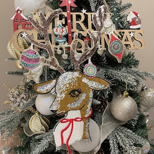 May include: A sparkly brown reindeer ornament with a red ribbon and white fur. The ornament is hanging on a Christmas tree with a wooden sign that says "Merry Christmas".