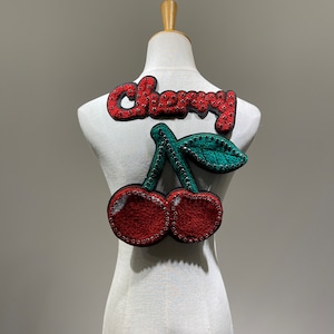 Può includere: Un manichino di stoffa bianca con un'applicazione di ciliegia in feltro rosso e verde e la parola "Cherry" in lettere di feltro rosso.