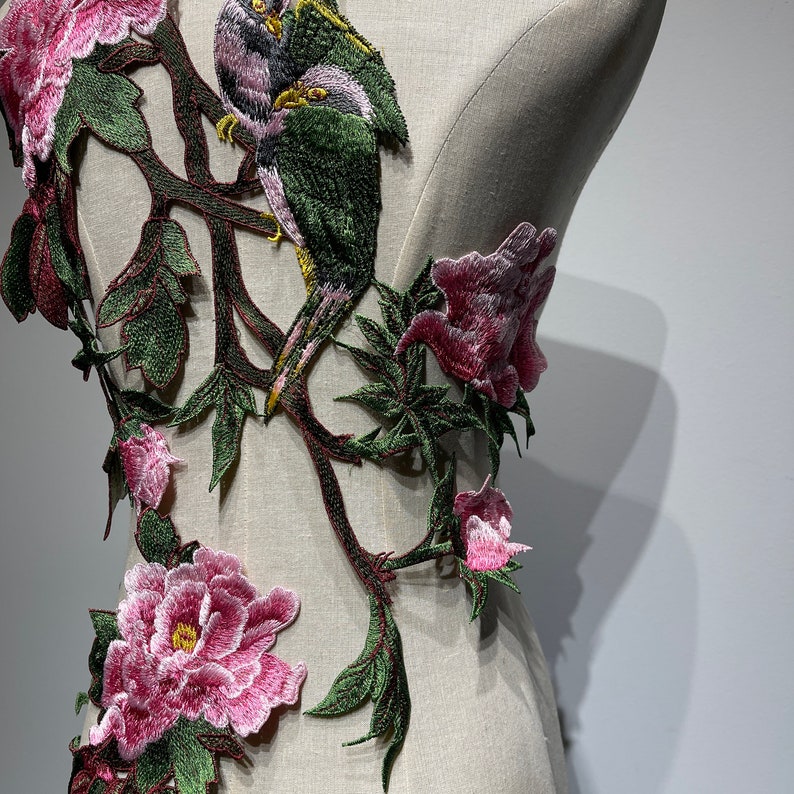 Pode incluir: Aplique bordado com um design floral com flores cor-de-rosa e folhas verdes. Dois p&aacute;ssaros est&atilde;o empoleirados num ramo, um verde e outro castanho.