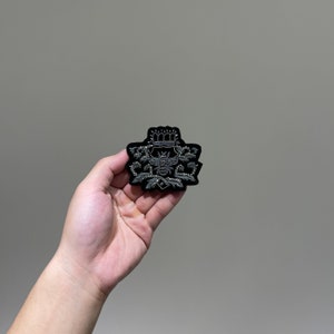 Può includere: Una toppa di velluto nero con un disegno ricamato in argento di un'ape con una corona sulla testa. L'ape è circondata da foglie e fiori.