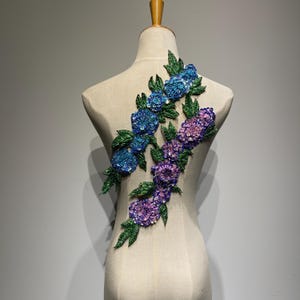 Könnte beinhalten: Eine perlenbesetzte Blumenapplikation mit blauen, violetten und grünen Akzenten. Die Applikation besteht aus Pailletten und Perlen und ist perfekt, um jedem Projekt einen Hauch von Glanz zu verleihen.