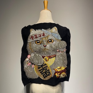 May include: Black fabric patch featuring an embroidered gray cat with large yellow eyes, a blue headband, and a gold coin. The cat is in a beckoning pose, holding a gold coin with Chinese characters. The patch is displayed on a mannequin.
