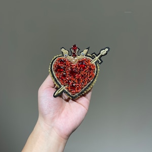Peut inclure: Un cœur rouge avec une couronne dorée et une épée qui le transperce. Le cœur est décoré de perles rouges et dorées.