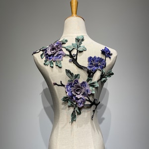 Könnte beinhalten: Eine zarte Blumenapplikation mit einem Zweig mit violetten und blauen Blüten und grünen Blättern. Die Applikation ist perfekt, um Kleidung, Accessoires oder Wohnaccessoires einen Hauch von Eleganz zu verleihen.