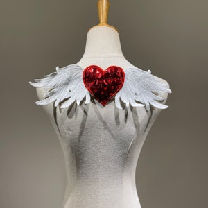 May include: A white fabric angel wing applique with a red sequined heart in the centre.