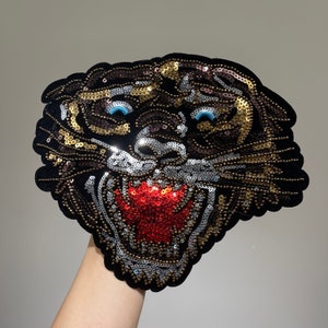 May include: A black velvet tiger head patch, embellished with sequins in gold, silver, and brown. The tiger has blue eyes and a red mouth, creating a striking contrast. This decorative patch is suitable for clothing and accessories.