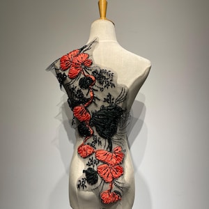 Peut inclure: Applique florale décorative avec des fleurs et des feuilles en tissu rouge et noir, ornée de perles noires. L'applique est fixée sur un support en résille transparente de couleur claire, présentée sur un mannequin.
