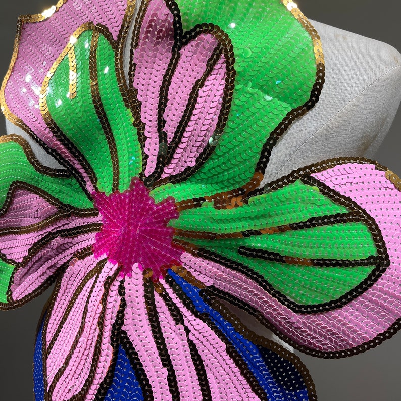May include: A large pink and green flower-shaped sequin applique with gold accents. The sequins are arranged in a pattern that resembles a flower with a pink center and green petals.