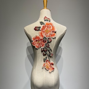 Könnte beinhalten: Gestickte Blumenapplikation mit orangefarbenen und roten Pfingstrosenblüten. Auf einer Schaufensterpuppe befestigt, zeigt sie das Potenzial zur Verschönerung von Kleidungsstücken. Die Blumen haben gelbe Zentren und grüne Blätter. Das Design ist sehr detailliert.