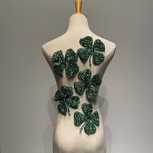May include: A white dress form with five green shamrock-shaped embellishments sewn onto the back. The shamrocks are made of sequins and beads.