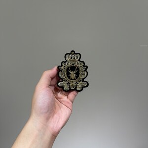 May include: A black velvet patch with gold embroidery depicting a crown and a spider in a circular frame.