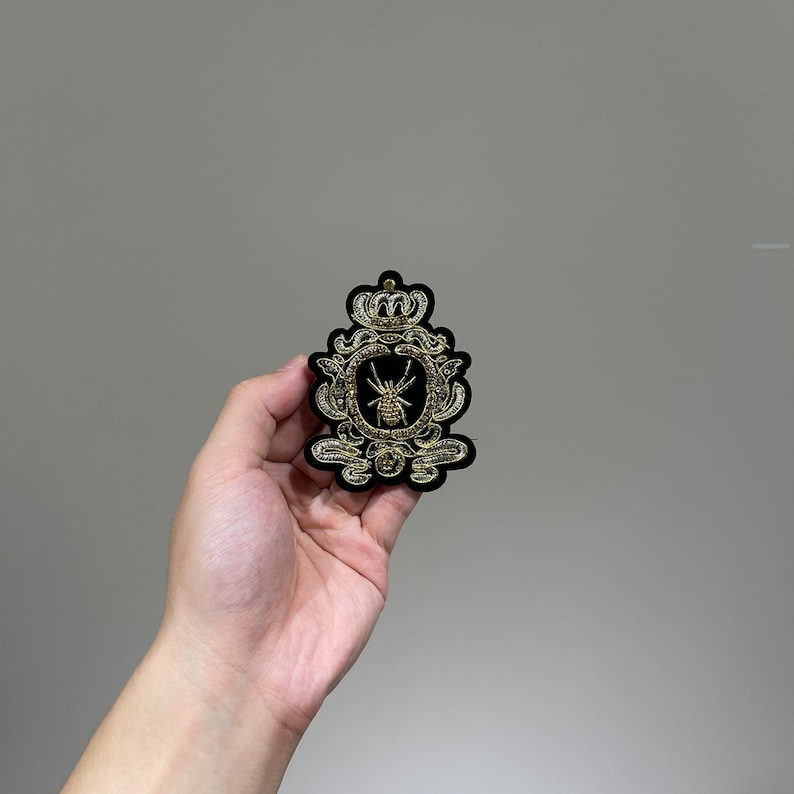 May include: A black velvet patch with gold embroidery depicting a crown and a spider in a circular frame.