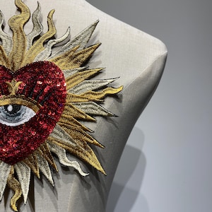 A red heart with a blue eye and eyelashes surrounded by a gold and silver sunburst design. The heart is made of sequins and the sunburst is embroidered.