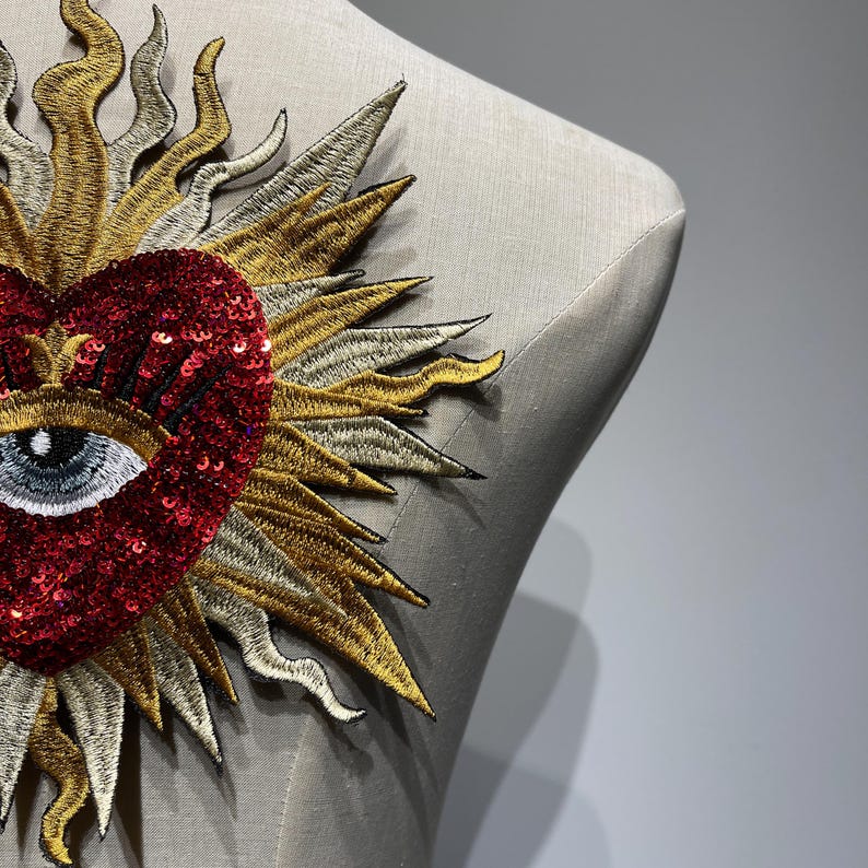 A red heart with a blue eye and eyelashes surrounded by a gold and silver sunburst design. The heart is made of sequins and the sunburst is embroidered.