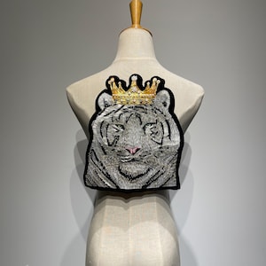 Pode incluir: Patch bordado com um tigre branco usando uma coroa de ouro. O tigre tem listras prateadas e pretas detalhadas, olhos azuis e um nariz rosa. O patch é contornado em preto.