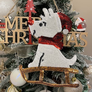 Pode incluir: Figura de cão branco com lantejoulas com um cachecol vermelho de lantejoulas a andar de trenó dourado com as palavras "Merry Christmas" num letreiro de madeira atrás.