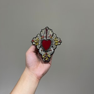 Può includere: Un cuore rosso con decorazioni dorate e argentate, circondato da un design a forma di croce. Il cuore è ricamato con perline e paillettes.