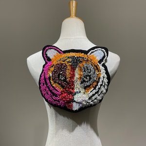 May include: A close-up of a tiger face applique made of colourful beads and sequins. The tiger's face is a mix of pink, orange, black, and white beads. The applique is attached to a white fabric background.
