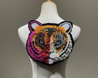 May include: A close-up of a tiger face applique made of colorful beads and sequins. The tiger's face is a mix of pink, orange, black, and white beads. The applique is attached to a white fabric background.