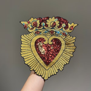 A gold and red embroidered heart with a crown on top. The heart is made of sequins and the crown is made of thread. The heart is surrounded by a gold embroidered halo.