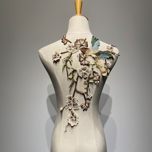 May include: A brown, green and white embroidered appliqué featuring a bird and floral design. The appliqué is on a white fabric background.