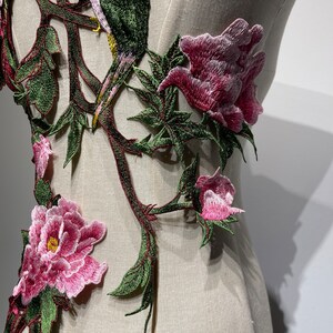 Pode incluir: Close-up de um desenho de bordado floral com flores cor-de-rosa e folhas verdes. O bordado est&aacute; sobre um fundo de tecido branco.