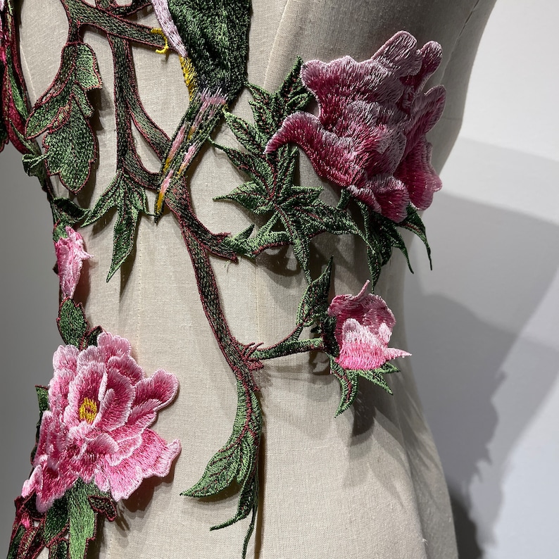 Pode incluir: Close-up de um desenho de bordado floral com flores cor-de-rosa e folhas verdes. O bordado est&aacute; sobre um fundo de tecido branco.