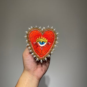 Può includere: Un broche ricamato a forma di cuore rosso con una corona dorata e un occhio blu con accenti turchesi e bianchi. Il cuore è circondato da perline di perle bianche.