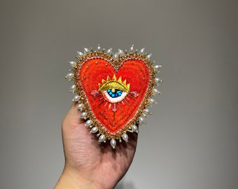May include: A red heart-shaped embroidered brooch with a gold crown and a blue eye with turquoise and white accents. The heart is surrounded by white pearl beads.