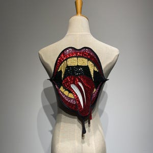 Op de afbeelding: Een grote, met pailletten bezette patch in de vorm van open lippen met hoektanden en een druipende tong. De lippen zijn rood en zwart, met gouden tanden en een zwart interieur. De tong is rood met witte accenten.
