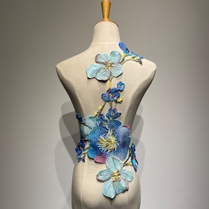 Könnte beinhalten: Eine blaue und grüne Blumenapplikation auf einem hellblauen Hintergrund. Die Applikation ist wie eine Rankenform gestaltet und zeigt mehrere Blumen und Blätter.