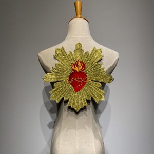 Può includere: Toppa ricamata del Sacro Cuore in oro e rosso. La toppa presenta un cuore rosso con una corona di spine, circondato da raggi dorati. Una fiamma sorge dalla parte superiore del cuore. La toppa è esposta su un manichino.