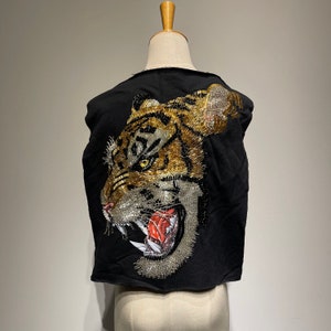 Pode incluir: Colete preto sem mangas com um grande design de rosto de tigre feito de lantejoulas douradas e prateadas. A boca do tigre está aberta e seus dentes são visíveis.