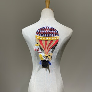May include: A red, white, and blue hot air balloon with a white and yellow striped pattern is embroidered on a white fabric. A white and yellow animal with a blue hat is holding onto the balloon basket.