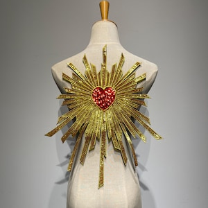 May include: A decorative applique featuring a central red heart covered in small, reflective red sequins. The heart is surrounded by a radiating sunburst pattern made of gold-colored, metallic-finish strips that extend outward in various lengths.