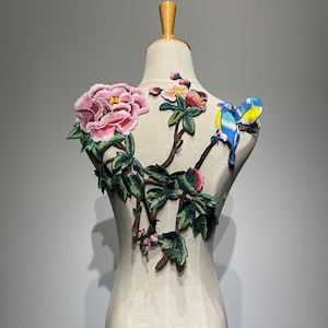 Puede incluir: Aplique bordado con una flor de peonía rosa, hojas verdes y dos pájaros azules con detalles amarillos. El aplique está sobre un fondo blanco.