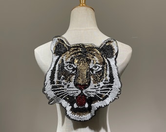 May include: A beaded tiger head patch with black, gold, white, and red accents. The tiger's face is intricately detailed with beads, showcasing its eyes, nose, and open mouth. The patch is designed to be sewn onto clothing or accessories.