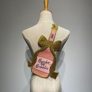 Może przedstawiać: Różowa i złota aplikacja z koralików w kształcie butelki szampana z napisem "Brooke of Bubbles". Aplikacja jest przymocowana do manekina. Butelka ma złotą kokardę i różową etykietę.